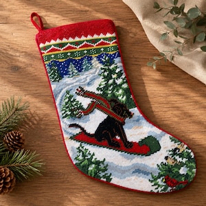 May include: A Christmas stocking featuring a needlepoint design of a black dog wearing a red scarf while sitting on a red sled in a snowy forest. The top border has a geometric pattern in red, green, and white, and a small red hanging loop.