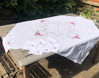 May include: A white tablecloth with embroidered floral designs and a set of matching napkins. The tablecloth features red and pink roses with green leaves, and a decorative border. The napkins have a small embroidered flower.