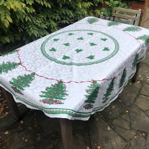 May include: White tablecloth with a red and green border featuring a repeating pattern of Christmas trees. The tablecloth has a scalloped edge and is laid out on a table.