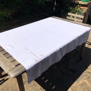 May include: White tablecloth with embroidered floral designs in shades of pink, blue, green, and yellow. The tablecloth is laid out on a wooden table.