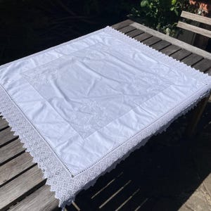 Könnte beinhalten: Quadratische weiße Tischdecke mit gestickten Blumendesigns in der Mitte und einem zarten Spitzensaum. Die Tischdecke besteht aus weichem, weißem Stoff und liegt auf einem Holztisch. Maße: ca. 150 cm x 150 cm.