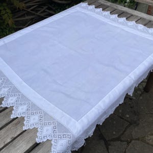 Op de afbeelding: Een witte tafelkleed met een decoratieve kanten rand. Het rechthoekige tafelkleed bedekt een houten tafel. De kanten rand is ingewikkeld en voegt een vleugje elegantie toe aan het eenvoudige ontwerp. Het tafelkleed is gemaakt van zachte, witte stof.