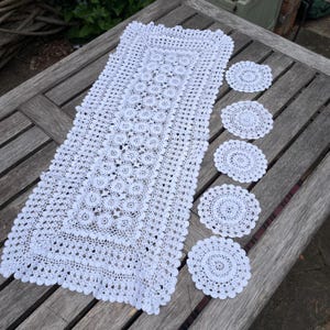 May include: A white, handmade crochet table runner and matching coasters. The runner is rectangular with a detailed floral pattern, and the coasters are round with a similar design. The set is displayed on a wooden table.