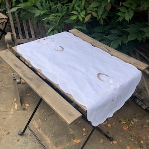 Può includere: Tovaglia da tavolo bianca ricamata con un bordo a festoni e motivi floreali. La tovaglia è appoggiata su un tavolo di legno.