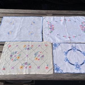 May include: Four rectangular fabric linens featuring various embroidery designs. One white cloth has blue floral motifs and a blue stitched border. Another white cloth displays pink and green floral garlands. A third cloth features a diamond pattern with colourful flowers. The fourth cloth has a blue floral wreath and corner accents.