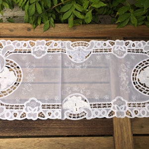 Puede incluir: Camino de mesa blanco bordado con motivos florales y geométricos. El camino de mesa presenta delicados calados y bordes festoneados. El diseño incluye flores, hojas y otros elementos decorativos. El camino de mesa está hecho de una tela fina y ligera.