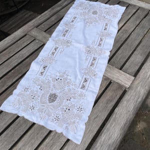 Puede incluir: Camino de mesa rectangular blanco con intrincados bordados florales y geométricos en beige. El camino de mesa, con borde festoneado, se exhibe sobre una mesa de madera, mostrando su diseño decorativo y artesanía.