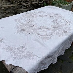 以下が含まれることがあります： 繊細な花の模様が刺繍された白いテーブルクロス。テーブルクロスは、波打つ縁取りが施され、軽量な生地で作られています。