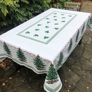 May include: White tablecloth with a green and red border featuring a repeating pattern of Christmas trees. The tablecloth is laid out on a table in an outdoor setting.