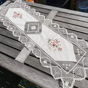 Puede incluir: Camino de mesa color crema con diseños florales bordados en rosa, melocotón y verde. El camino de mesa presenta un delicado borde de encaje y un cuadrado central con un motivo floral. Colocado sobre una mesa de madera.