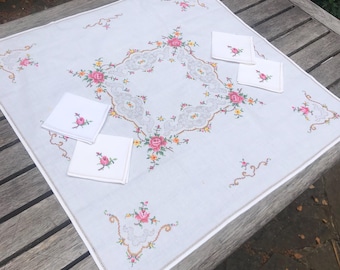 Può includere: Una tovaglia bianca ricamata con un motivo centrale a forma di diamante e accenti floreali rosa, gialli e verdi. Include quattro tovaglioli quadrati abbinati, ciascuno con una rosa. La tovaglia è su una superficie di legno.