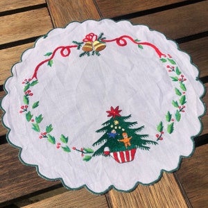 Peut inclure: Un chemin de table de Noël en lin blanc avec du houx brodé en vert et rouge, un sapin de Noël avec des ornements et deux enfants, et un ruban rouge avec deux cloches dorées.