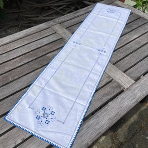Puede incluir: Camino de mesa rectangular blanco con diseños florales bordados en azul y un borde azul en zigzag. El camino de mesa está colocado sobre una mesa de madera desgastada, mostrando sus elementos decorativos y artesanía. Mide aproximadamente 30 cm de ancho y 122 cm de largo.
