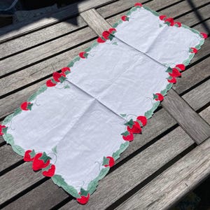 May include: White rectangular table runner with a scalloped edge. The border is decorated with red strawberries and green leaves. The runner is folded in thirds and placed on a wooden surface.