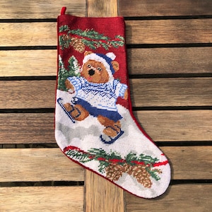 May include: A red and white Christmas stocking with a needlepoint design of a bear ice skating. The bear is wearing a blue and white sweater and a blue hat. The stocking is decorated with pine branches and pine cones.