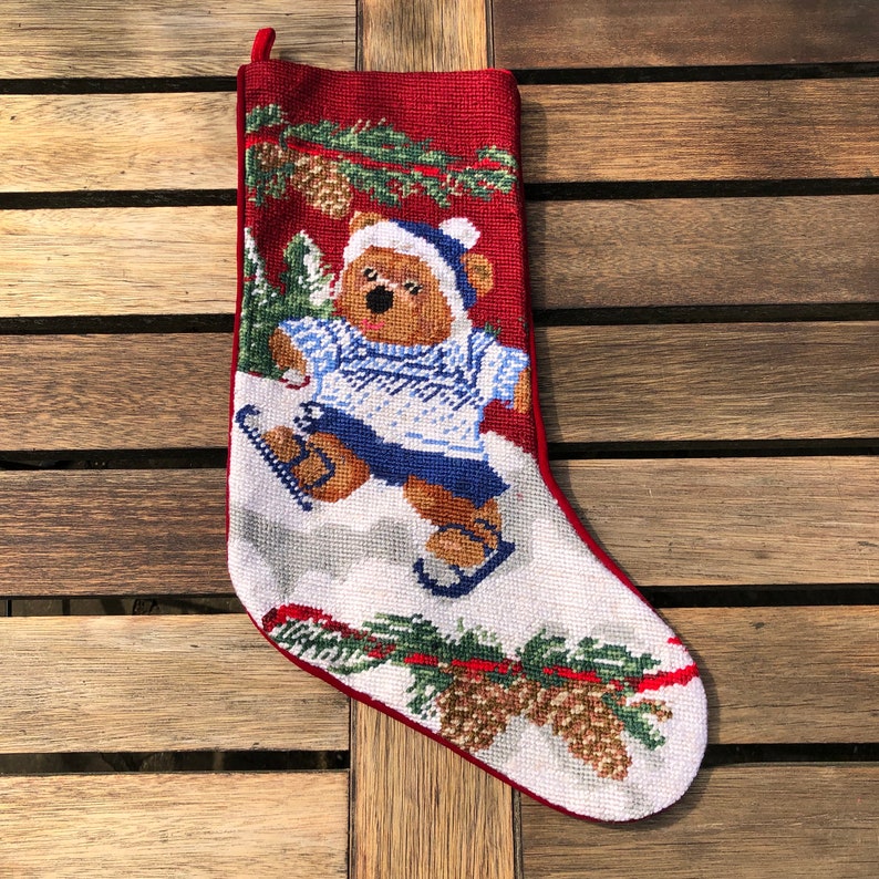 May include: A red and white Christmas stocking with a needlepoint design of a bear ice skating. The bear is wearing a blue and white sweater and a blue hat. The stocking is decorated with pine branches and pine cones.