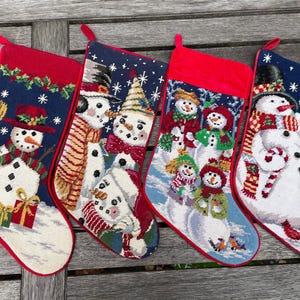 Puede incluir: Cuatro calcetines navideños con ribete rojo y presillas para colgar. Cada calcetín presenta un diseño de muñeco de nieve diferente sobre un fondo azul con copos de nieve. Los muñecos de nieve llevan sombreros, bufandas y sostienen regalos y bastones de caramelo.