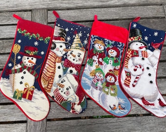 Puede incluir: Cuatro calcetines navideños con ribete rojo y presillas para colgar. Cada calcetín presenta un diseño de muñeco de nieve diferente sobre un fondo azul con copos de nieve. Los muñecos de nieve llevan sombreros, bufandas y sostienen regalos y bastones de caramelo.
