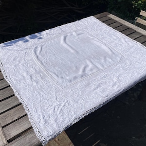 Op de afbeelding: Een witte, rechthoekige tafelkleed met ingewikkelde bloemenborduurwerk en een delicate gehaakte rand. Het tafelkleed ligt op een houten tafel en toont het gedetailleerde ontwerp en de textuur. Het centrale gedeelte is effen.