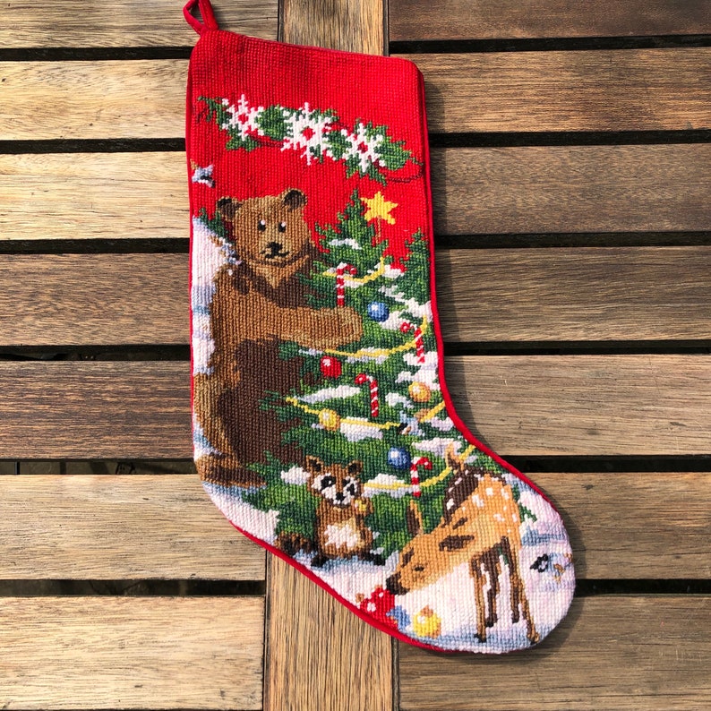 May include: Red Christmas stocking with a white background featuring a brown bear, a raccoon, and a deer standing around a green Christmas tree with red and white candy canes, ornaments, and lights.
