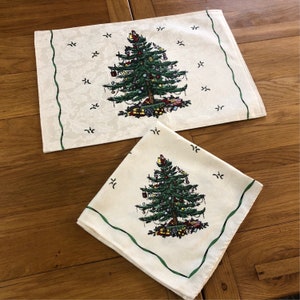 Op de afbeelding: Twee witte katoenen placemats met een groene rand en een geprinte afbeelding van een kerstboom met ornamenten en cadeaus. De placemats liggen op een houten oppervlak.