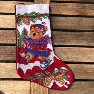 May include: Red and white Christmas stocking with a brown bear wearing a blue sweater and ice skating on a snowy background. The stocking is decorated with pine branches and pine cones.