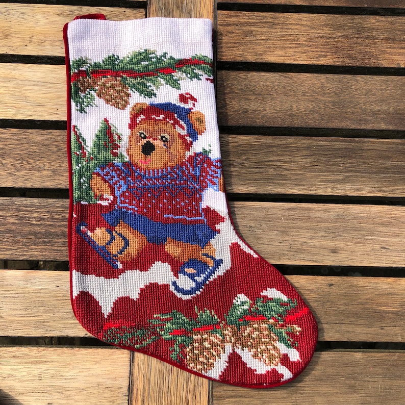 May include: Red and white Christmas stocking with a brown bear wearing a blue sweater and ice skating on a snowy background. The stocking is decorated with pine branches and pine cones.