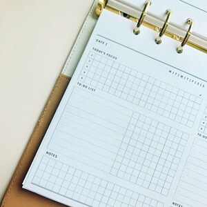 May include: A white planner with a gold binder. The planner has a grid layout with sections for "Date", "Today's Focus", "To-Do List", and "Notes". The days of the week are labeled "M", "T", "W", "Th", "F", "S", and "S".