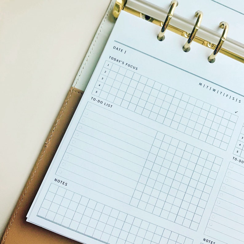 May include: A white planner with a gold binder. The planner has a grid layout with sections for "Date", "Today's Focus", "To-Do List", and "Notes". The days of the week are labeled "M", "T", "W", "Th", "F", "S", and "S".