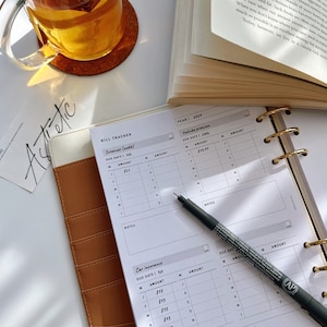 May include: A white planner with a brown leather cover. The planner has a bill tracker section with columns for due date, amount, and notes. The planner also has a section for car insurance with columns for due date, amount, and notes. The planner is open to the bill tracker section and a black pen is lying on top of the page.