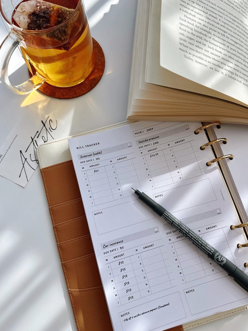 May include: A white planner with a brown leather cover. The planner has a bill tracker section with columns for due date, amount, and notes. The planner also has a section for car insurance with columns for due date, amount, and notes. The planner is open to the bill tracker section and a black pen is lying on top of the page.