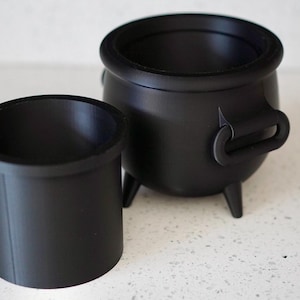 Pode incluir: Dois caldeirões pretos, um pequeno e cilíndrico, o outro maior com corpo arredondado, três pernas e uma alça. Os caldeirões são feitos de um material preto fosco e provavelmente são usados para fins decorativos ou de novidade.