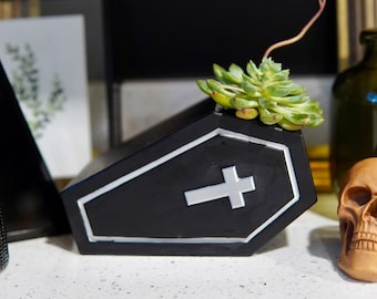 May include: A black coffin-shaped planter with a white cross on the front. The planter is filled with a green succulent plant. A small, brown skull figurine is in the foreground.