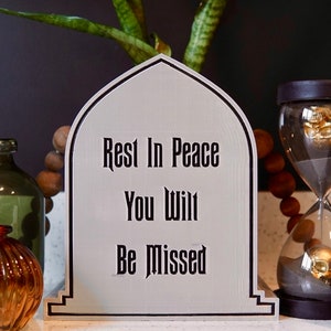 May include: A white tombstone with black lettering that reads "Rest In Peace You Will Be Missed". The tombstone is surrounded by decorative items, including a glass hourglass with a gold skull inside, a green glass bottle, a brown glass bottle, and a beaded garland.