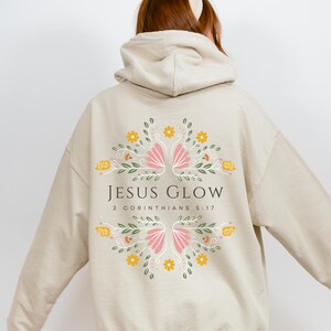 May include: A beige hoodie with a floral design and the text "JESUS GLOW 2 CORINTHIANS 5:17" printed on the back.