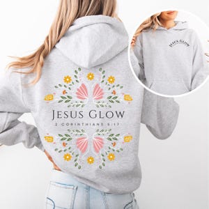 May include: Gray hooded sweatshirt with a floral design on the back. The design features pink flowers, yellow flowers, and green leaves. The text "JESUS GLOW" and "2 CORINTHIANS 5:17" is printed on the back of the sweatshirt.