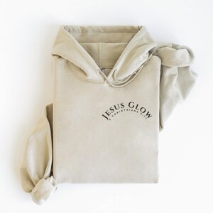 May include: A beige hoodie with the text "JESUS GLOW 2 CORINTHIANS 1:20" printed on the front.