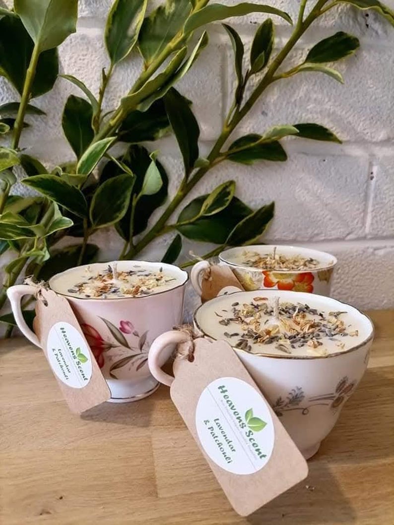 May include: Three vintage teacups filled with soy wax candles. The candles are topped with dried flowers and herbs. The teacups are decorated with floral patterns. The candles are labeled "Heavens Scent" and "Lavender & Patchouli".