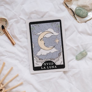Pode incluir: Um cart&atilde;o de tar&ocirc; com uma borda preta e uma lua crescente branca com um rosto nela. O cart&atilde;o est&aacute; sobre um fundo branco com uma estrela e nuvens. O texto no cart&atilde;o diz "XVIII - LA LUNA".