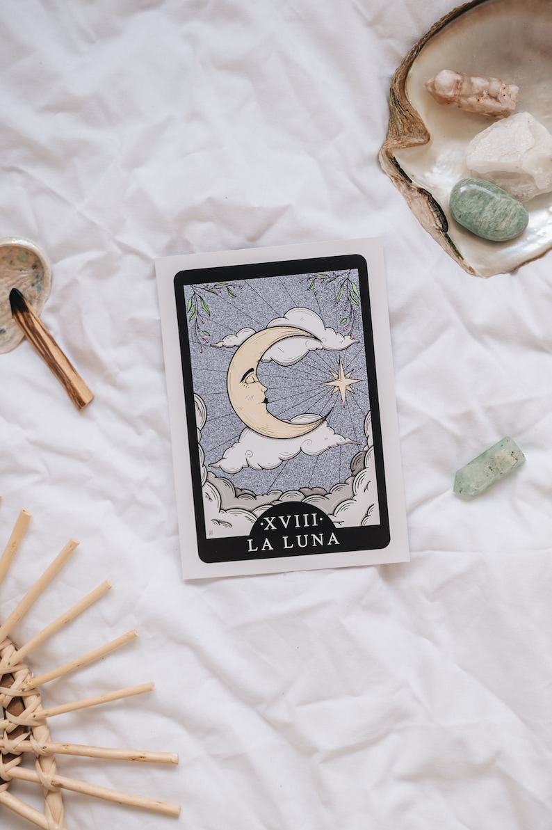 Pode incluir: Um cart&atilde;o de tar&ocirc; com uma borda preta e uma lua crescente branca com um rosto nela. O cart&atilde;o est&aacute; sobre um fundo branco com uma estrela e nuvens. O texto no cart&atilde;o diz "XVIII - LA LUNA".