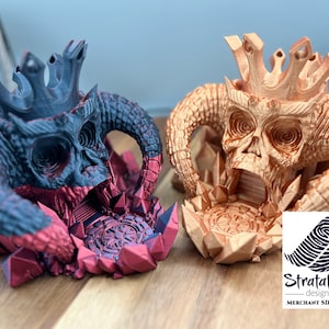 May include: Two 3D printed skull dice towers, one in black and red and the other in orange. Both towers feature a crown and horns. The towers are on a wooden surface. Strataion Design - Merchant SD-0256