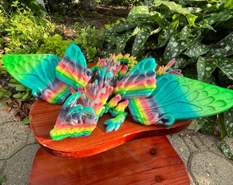 May include: A multi-colored, glittery, 3D printed dragon sculpture with butterfly wings. The dragon is made of several pieces and is laying on a wooden surface.