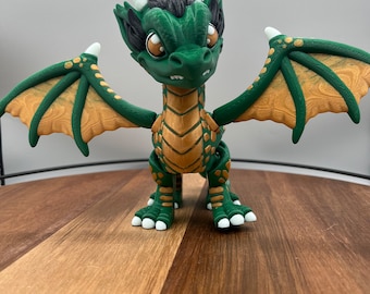 May include: A green and gold dragon figurine with outstretched wings. The dragon has large, expressive eyes, white horns, and a spiky crest. The wings are detailed with a gold pattern. The figurine is posed on a wooden surface.