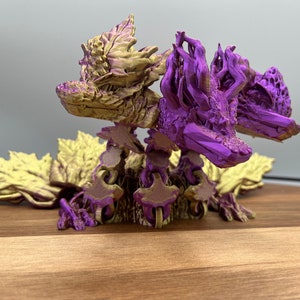 May include: Two 3D printed dragons, one gold and one purple, are posed on a wooden surface. The dragons are intricately detailed and have a leafy, organic design.