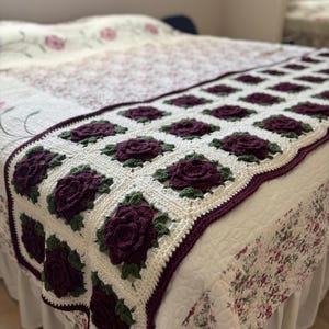 May include: A handmade crochet blanket featuring a repeating pattern of burgundy roses with green leaves on a white background. The blanket has a burgundy border and is placed on a bed with a floral patterned quilt and bed skirt.