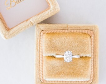 May include: A ring with a large, round, clear diamond is displayed in a small, square, tan velvet ring box. The box is open and the ring is sitting on the velvet inside. The box has the word "Balacia" written on the inside lid.