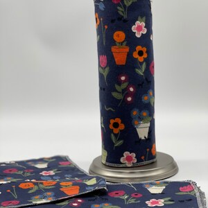 Könnte beinhalten: Ein marineblauer Papiertuchhalter mit einem Blumenmuster mit orangefarbenen, rosa und weißen Blumen in Töpfen. Zwei passende Tücher befinden sich im Vordergrund. Der Halter steht auf einer silbernen Basis.