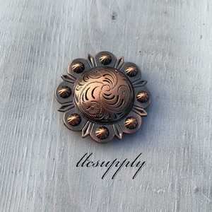 May include: A copper-toned metal concho with an intricate floral design. The concho has a circular shape with a raised center and a decorative border.