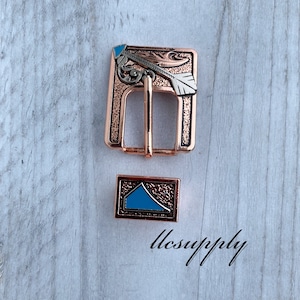 May include: A copper-colored metal belt buckle and matching keeper with an arrow design and turquoise accents. The buckle is rectangular with a decorative arrow and leaf design. The keeper is a small rectangle with a turquoise design. The text "llsupply" is visible.