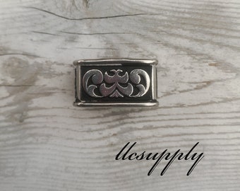 May include: A silver metal buckle with an ornate design. The buckle is rectangular and has a black background.